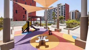 A playground with a slide and swings, surrounded by buildings and shaded by large canopies. - PVH - Azizi Riviera 37 Vibrant Studio (Dubai)