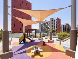 A playground with a slide and swings, surrounded by buildings and shaded by large canopies. - PVH - Azizi Riviera 37 Vibrant Studio (Dubai)