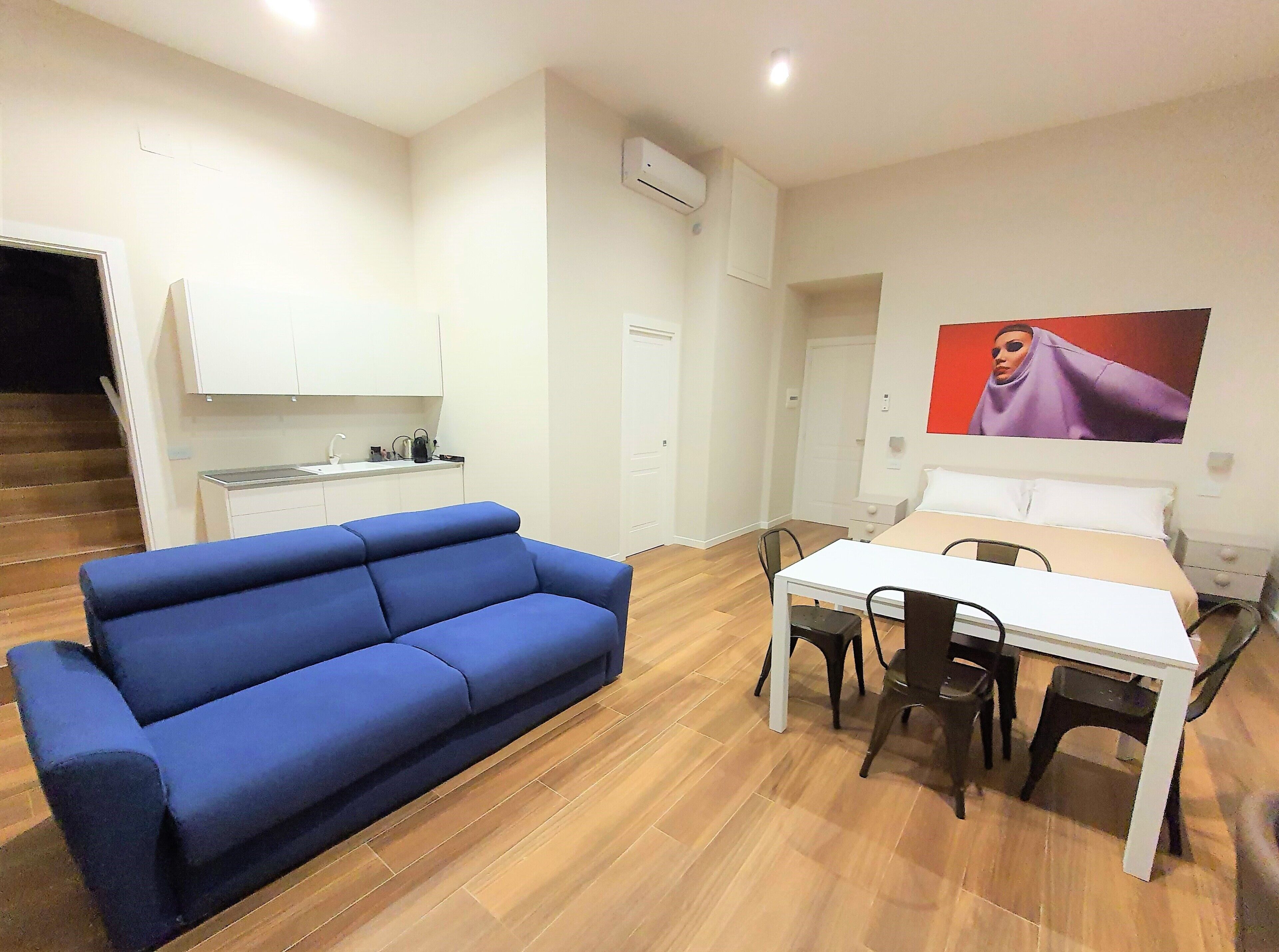Ein Zimmer mit blauem Sofa, einem Esstisch mit Stühlen, einem Bett, einer Küchenzeile und einem Bild an der Wand.