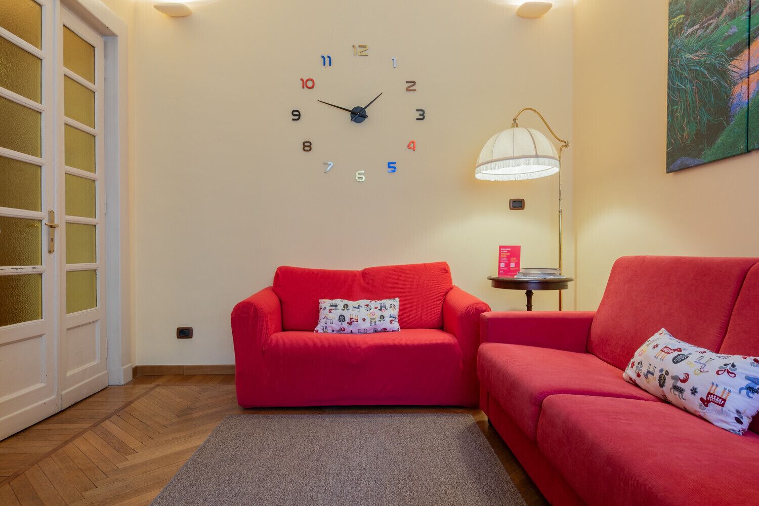 Ein Zimmer mit einem roten Sofa, einer Wanduhr, einer Lampe und einer Glastür.