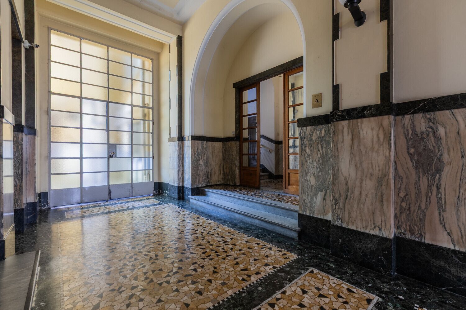 A hallway with a tiled floor, a door with frosted glass panels, and a staircase with a marble finish.