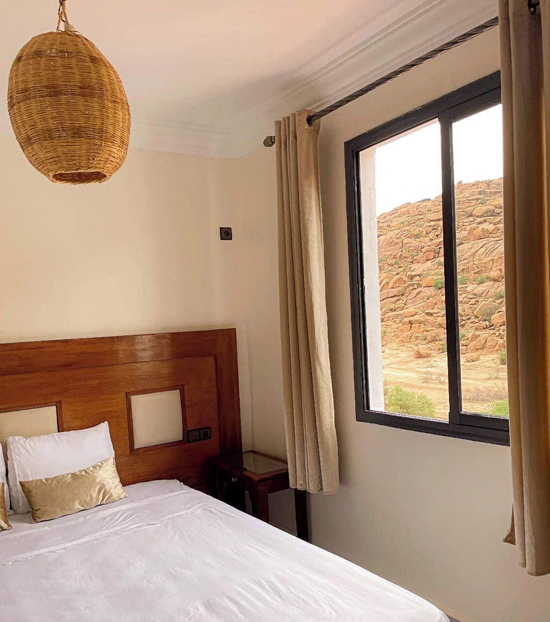 Habitación de hotel con cabecera de madera, cama grande con ropa blanca, mesita de noche, lámpara colgante de mimbre y ventana con cortinas.