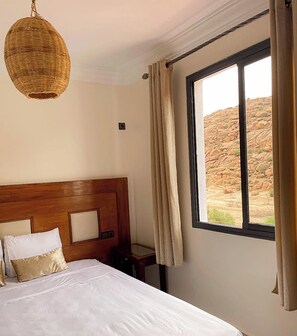 Habitación de hotel con cabecera de madera, cama grande con ropa blanca, mesita de noche, lámpara colgante de mimbre y ventana con cortinas.