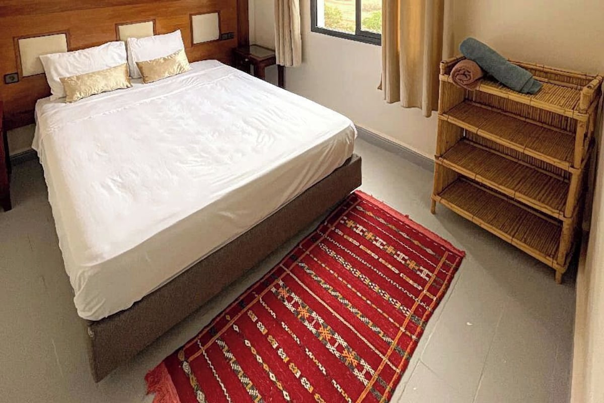Ein Schlafzimmer mit einem großen Bett, einem Nachttisch, einem Fenster mit Vorhängen, einem gewebten Korb und einem roten Teppich mit Muster.