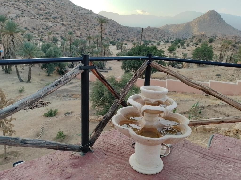A terracotta water fountain with multiple tiers, set against a backdrop of a desert landscape with sparse vegetation and distant mountains.