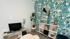 A modern living room with a flat-screen TV, a small table, two chairs, a bookshelf, and a potted plant.