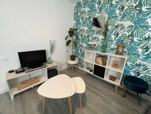 A modern living room with a flat-screen TV, a small table, two chairs, a bookshelf, and a potted plant.