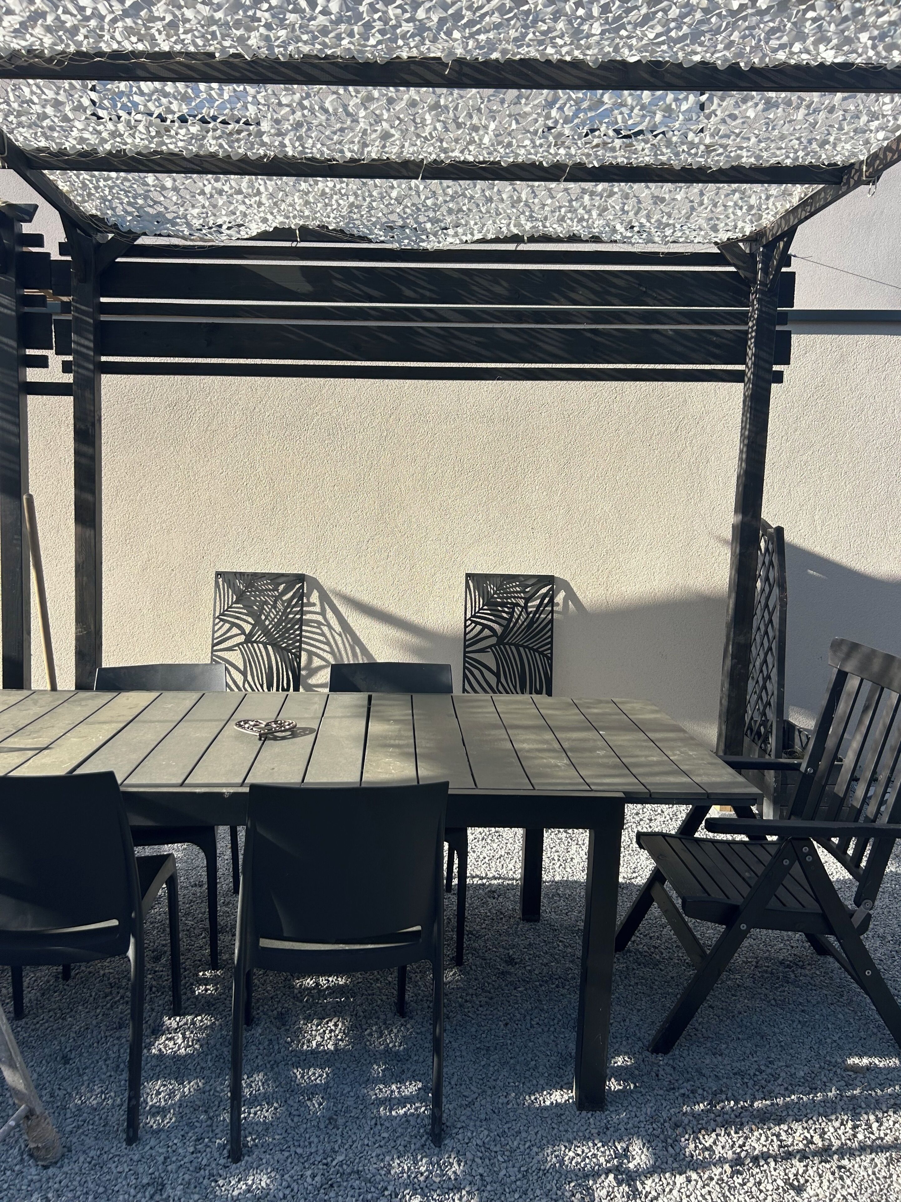 An outdoor patio with a wooden table and chairs, a pergola overhead, and a textured wall in the background.