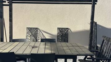 An outdoor patio with a wooden table and chairs, a pergola overhead, and a textured wall in the background.