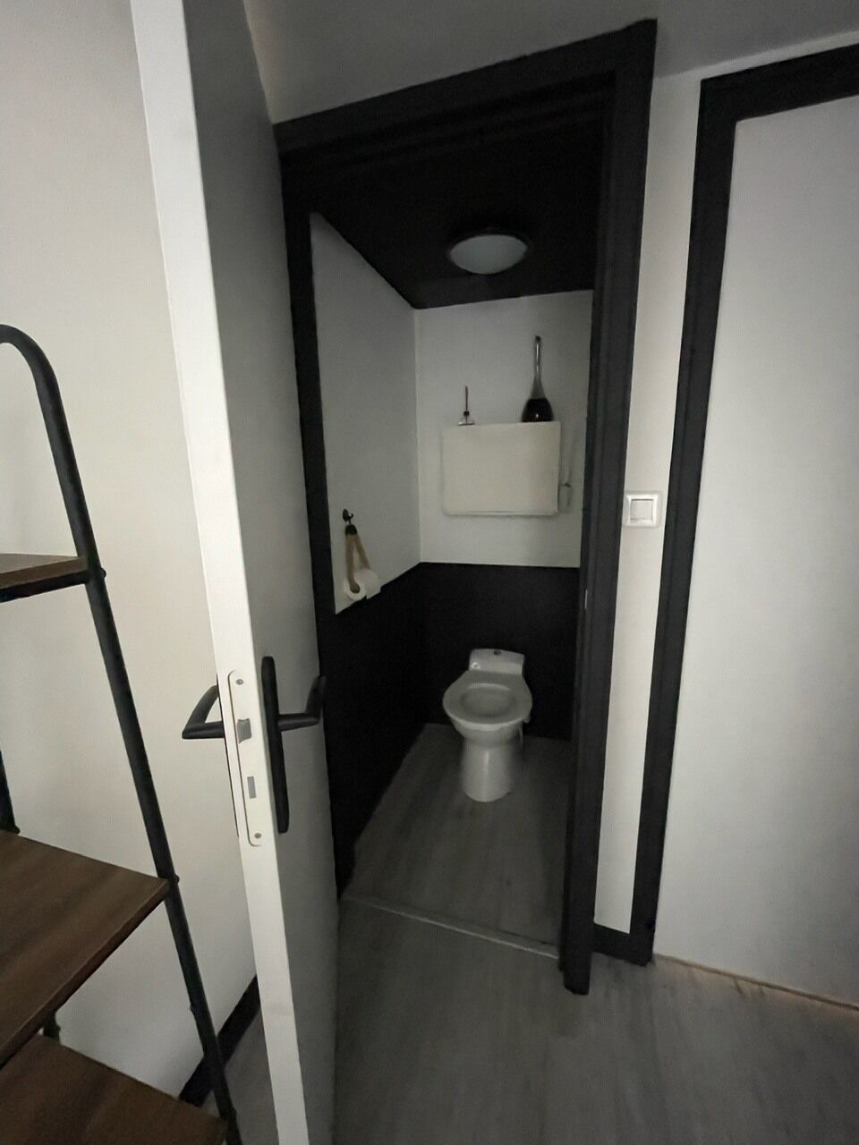Un couloir étroit mène à une salle de bain équipée de toilettes, d’une étagère sur laquelle se trouve une bouteille, et d’une serviette accrochée au mur.