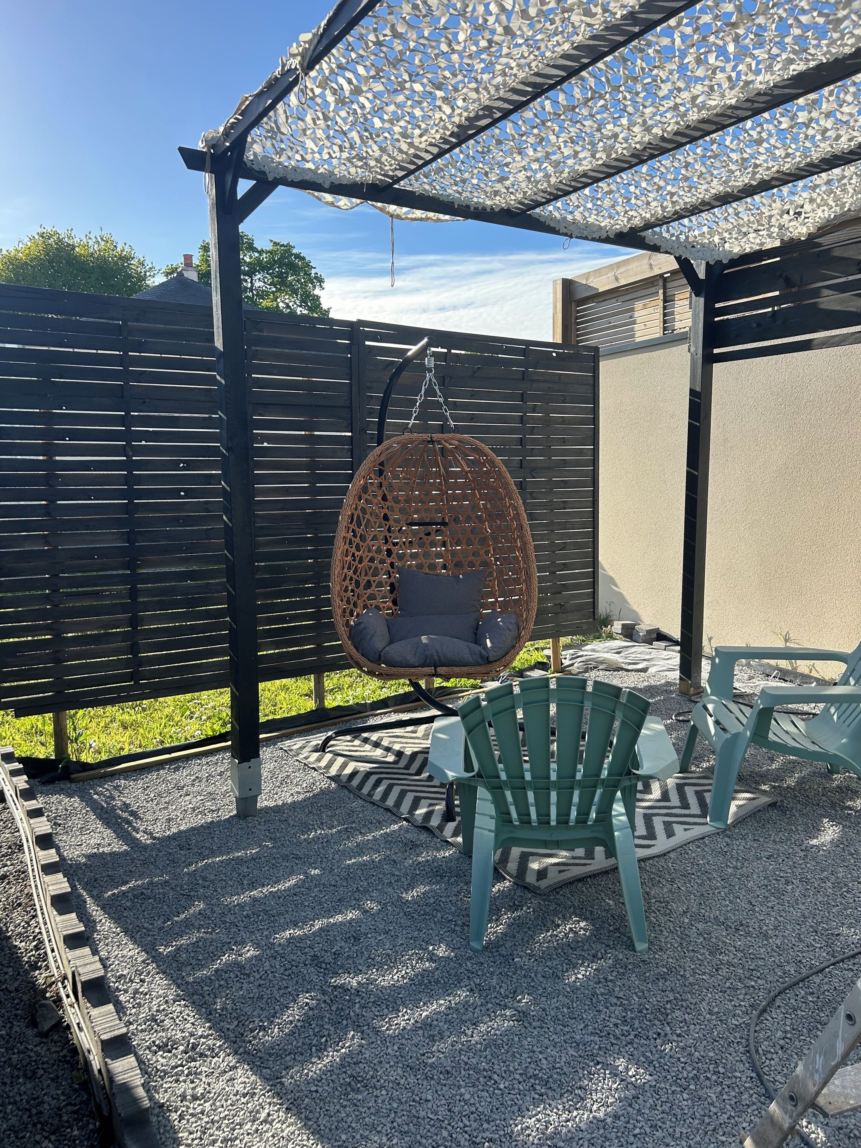 Een terras met een hangstoel van riet, groene plastic stoelen en een kamerkleed met dessin.