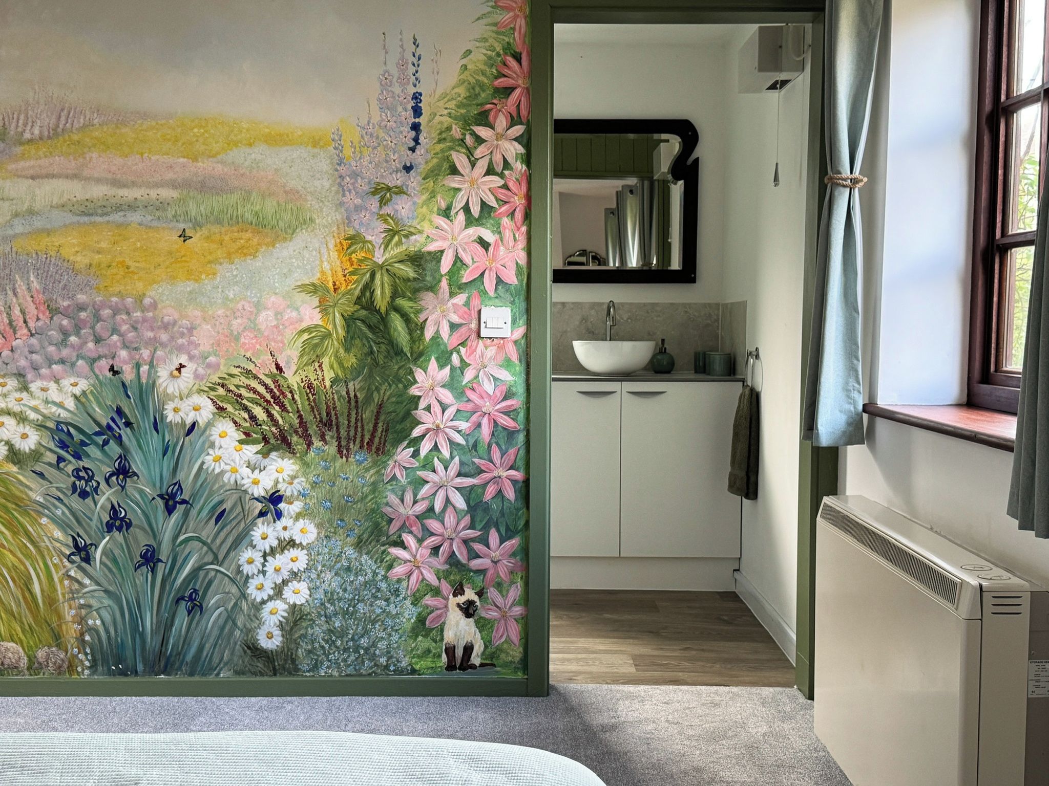 Une pièce agrémentée d’une fresque murale aux motifs floraux éclatants, d’une fenêtre avec des rideaux, d’un lavabo et d’un radiateur.