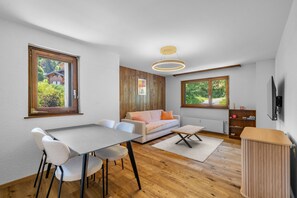 Ein modernes Wohnzimmer mit Essbereich, Holzboden, einer Couch, einem Couchtisch und einem an der Wand befestigten Fernseher.