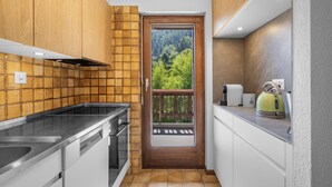 Uma cozinha compacta com vista para uma floresta através de uma porta de vidro, um fogão e uma chaleira sobre a bancada.