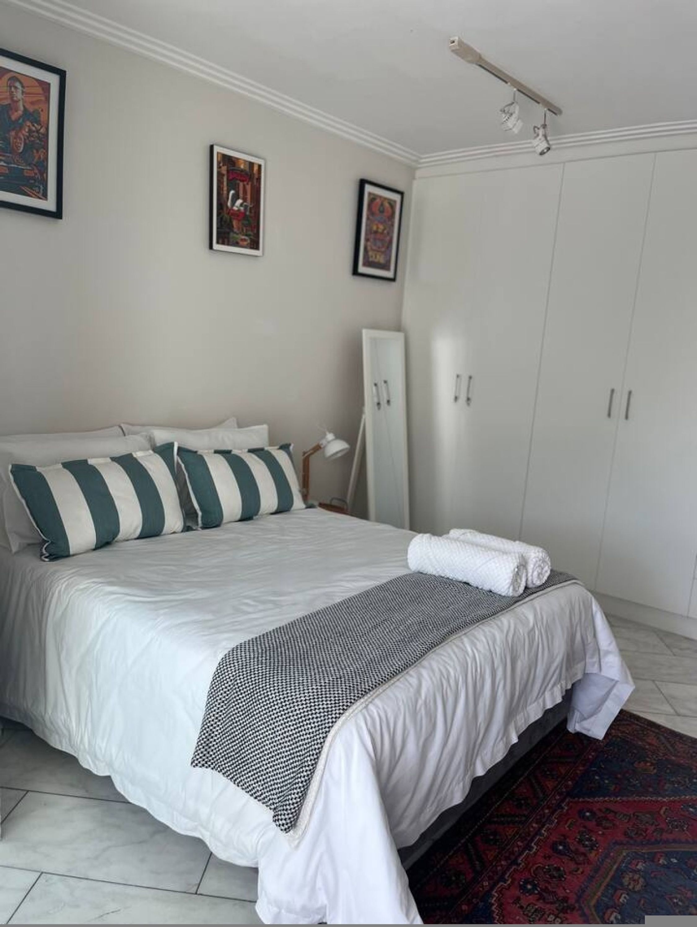 Una cama bien hecha con almohadones a rayas, una mesita de noche blanca con lámpara y un amplio armario con varias puertas.