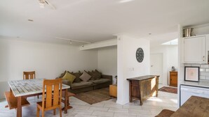 A dining area with a wooden table and chairs, a sofa with cushions, a clock on the wall, and a kitchen area with cabinets and a dishwasher. - Beachfront Designer Villa in Secure Estate (Cape Town)