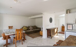 Ein Essbereich mit Holztisch und Stühlen, ein Sofa mit Kissen, eine Wanduhr und eine Küchenzeile mit Schränken und einem Geschirrspüler.