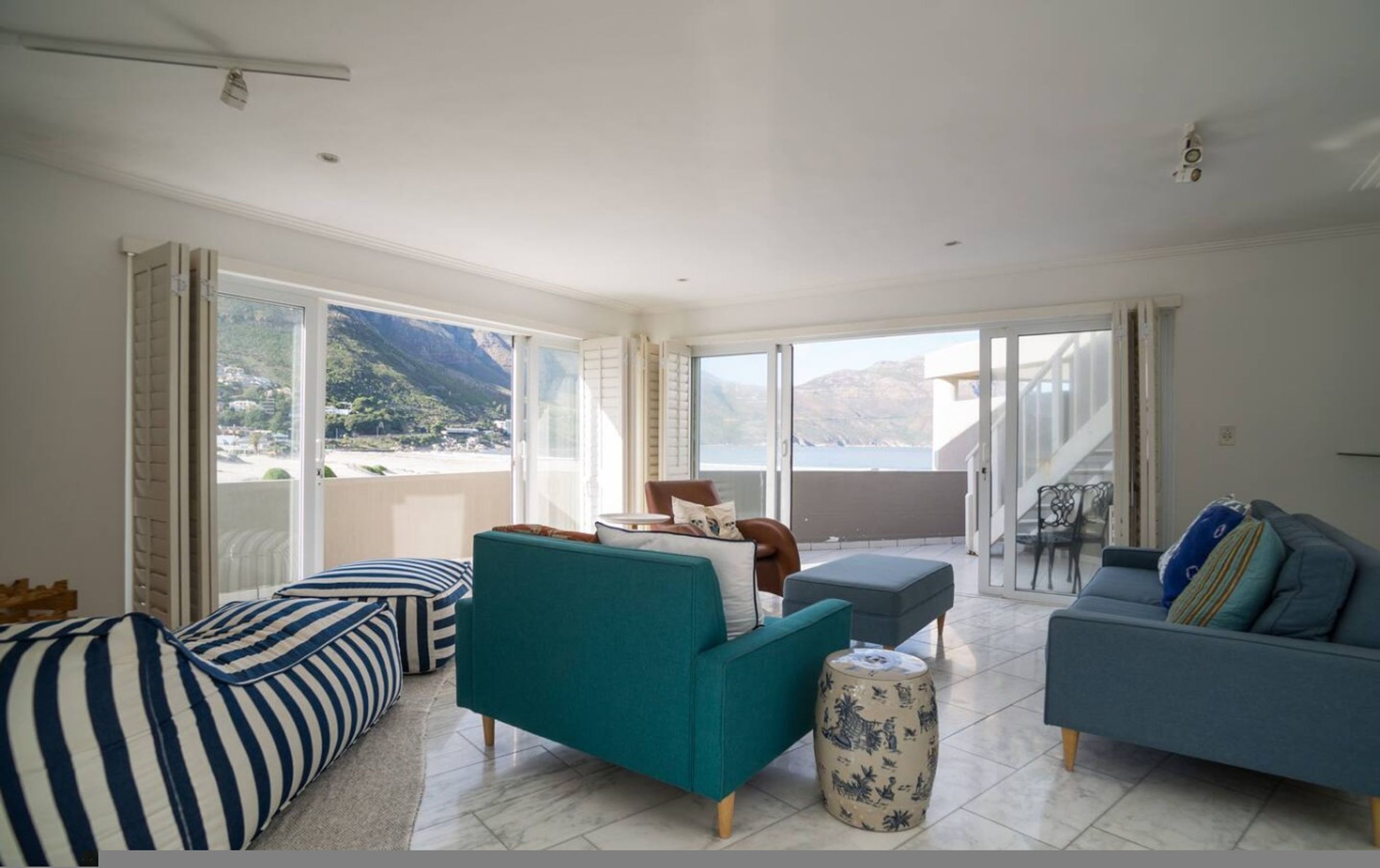 Amplia sala de estar con sofá, sillón y reposapiés, un balcón con vistas al mar y un comedor con mesa y sillas.