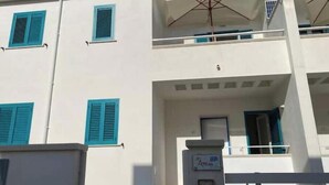 Un immeuble à plusieurs étages avec des murs blancs et des volets bleus, un balcon avec un parasol blanc, et une enseigne indiquant un commerce ou un espace de bureaux.