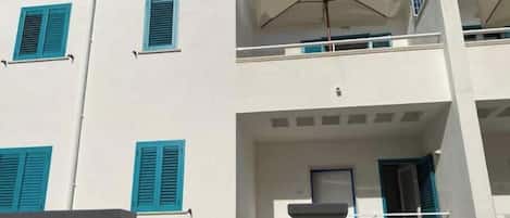 Ein mehrstöckiges Gebäude mit weißen Wänden und blauen Fensterläden, ein Balkon mit einem weißen Sonnenschirm und ein Schild, das auf Geschäfts- oder Büroflächen hinweist.