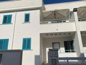 A multi-story building with white walls and blue shutters, a balcony with a white umbrella, and a sign indicating a business or office space. - Amida App.to, Cielo, 1st Floor (Campomarino di Maruggio (TA))