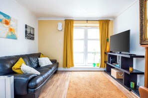 Ein Wohnzimmer mit einem schwarzen Ledersofa, gelben Kissen, einem Flachbildfernseher und Holzboden.