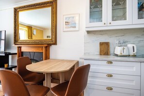Ein Essbereich mit einem Holztisch und braunen Stühlen, ein großer Spiegel, ein gerahmtes Bild sowie eine Küche mit weißen Schränken und einer Marmorarbeitsplatte.