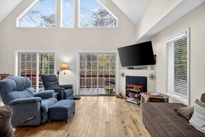 A spacious living room with a high ceiling, large windows, a flat-screen TV mounted on the wall, and a fireplace. - Spacious Woodland Cabin with Fire Pit & Creek Near Berkeley Springs, West Virginia (Berkeley Springs)