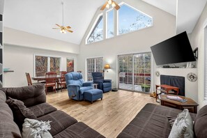 A spacious living room with a high ceiling, a ceiling fan, a flat-screen TV mounted on the wall, and a large sliding glass door leading to an outdoor area. - Spacious Woodland Cabin with Fire Pit & Creek Near Berkeley Springs, West Virginia (Berkeley Springs)