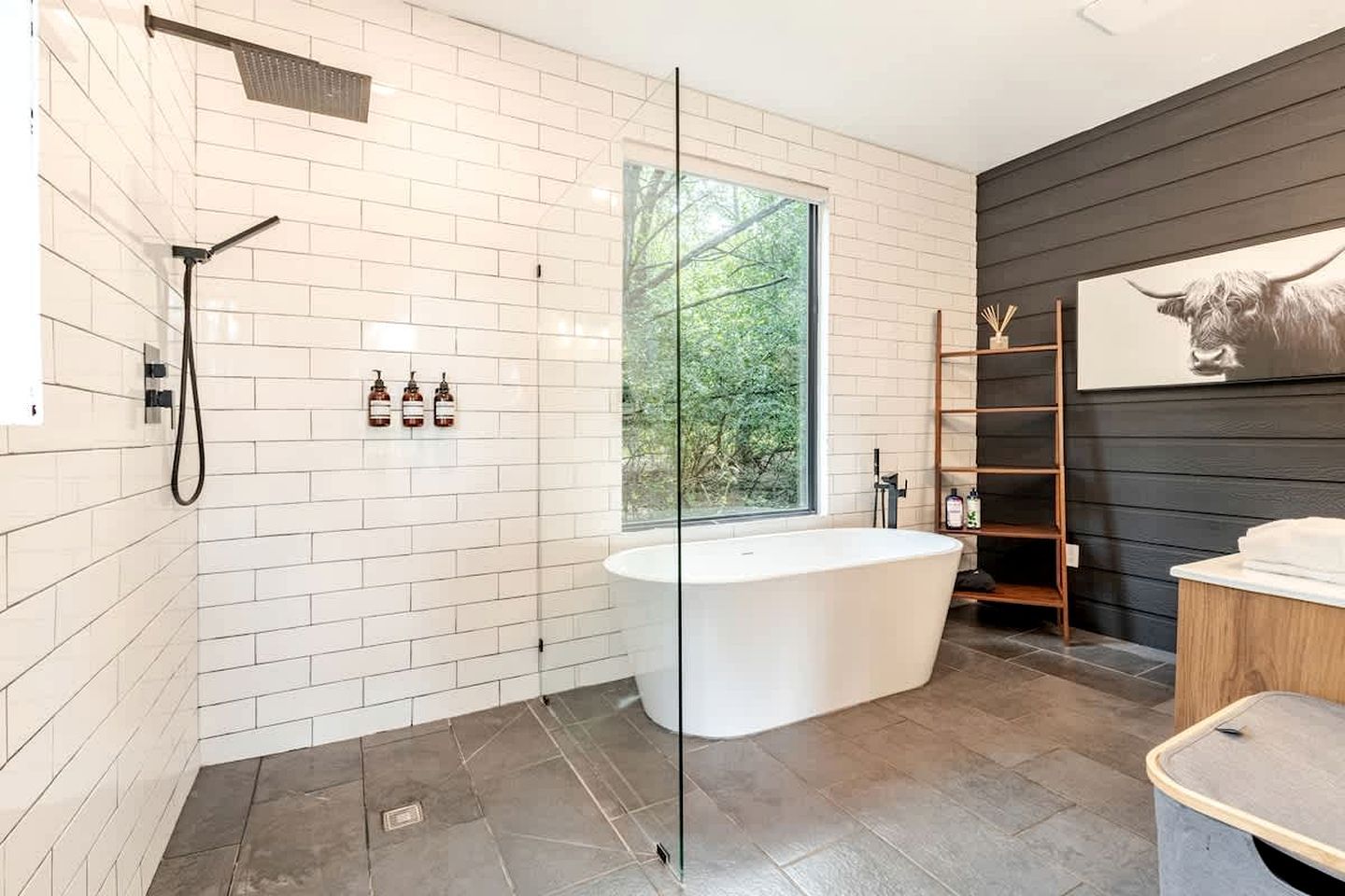 Ein modernes Badezimmer mit freistehender Badewanne, einer Duschkabine aus Glas und einem großen Fenster mit Blick auf Grünflächen.