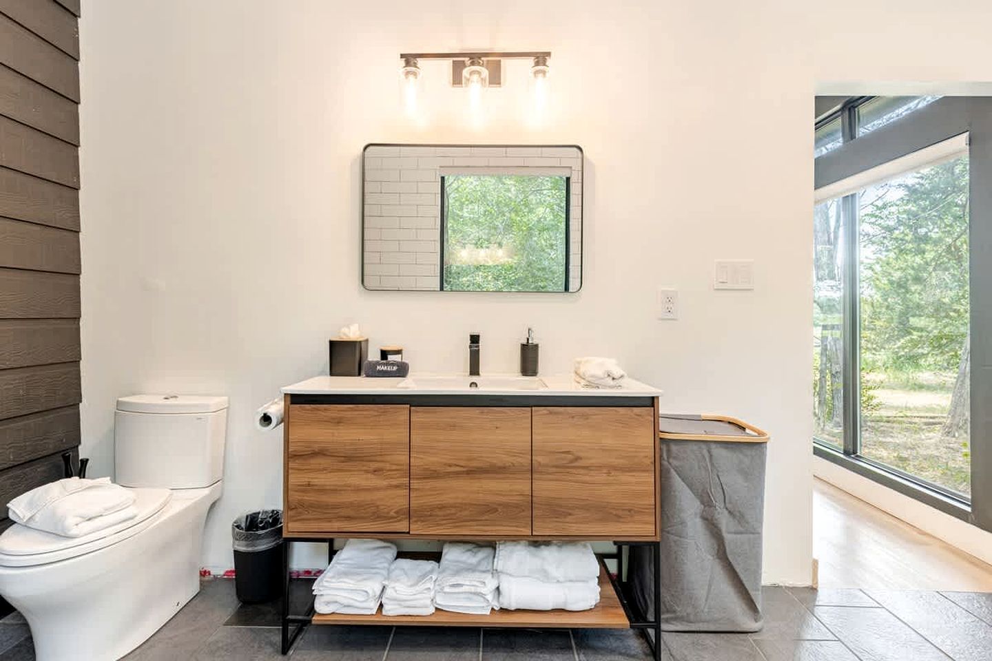 A modern bathroom with a wooden vanity, a toilet, and a large window providing a view of greenery.