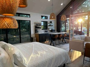 A modern bedroom with a large bed, a dining area with chairs, and a kitchen in the background.