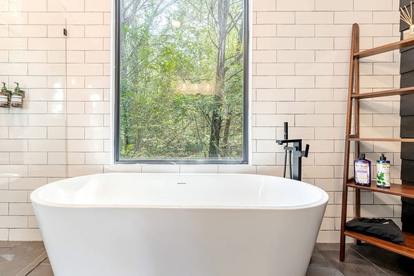 Un baño moderno con una bañera independiente, un ventanal con vistas a árboles y un estante de madera con artículos de aseo.
