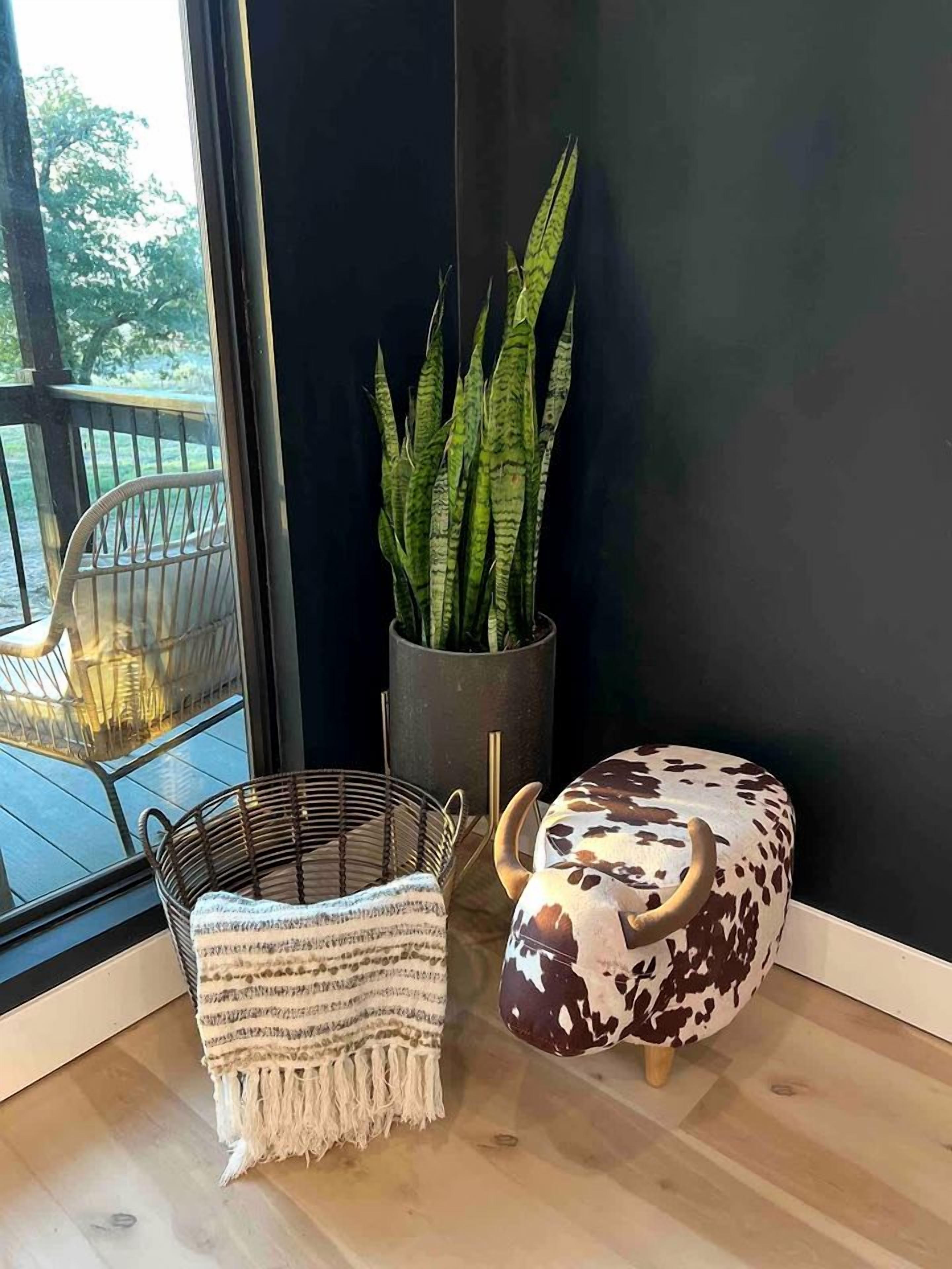 Une pièce dotée d'une grande fenêtre, d'un fauteuil en osier, d'un petit tabouret en fibres tressées, d'une plante en pot et d'une statue de taureau décorative.