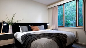 Une chambre à coucher comprenant un lit, des tables de chevet, une commode, une lampe, une fenêtre avec des rideaux et une vue sur la verdure à l’extérieur.
