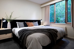 A bedroom with a bed, bedside tables, a nightstand, a lamp, a window with curtains, and a view of greenery outside. - Amazing Villa with Incredible Views and Outdoor Living Space in Daylesford, Victoria (Daylesford)