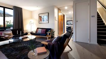 Un salon moderne avec un canapé, une table basse et un fauteuil. Un escalier se trouve sur la droite, et une porte donne accès à une autre pièce.