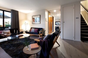 Un salon moderne avec un canapé, une table basse et un fauteuil. Un escalier se trouve sur la droite, et une porte donne accès à une autre pièce.