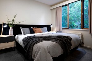 A bedroom with a bed, bedside tables, a nightstand, a lamp, a window with curtains, and a view of greenery outside.
