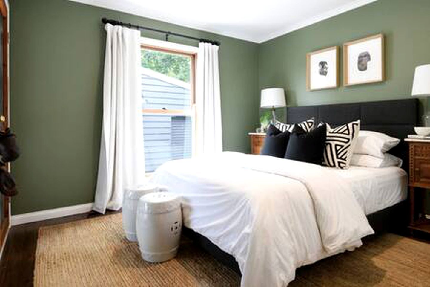 A bedroom with a bed, a nightstand, a lamp, a window with curtains, and a wall with two framed pictures.