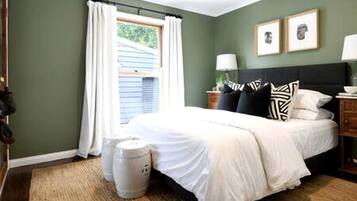 A bedroom with a bed, a nightstand, a lamp, a window with curtains, and a wall with two framed pictures.