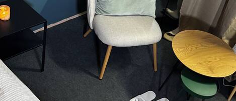 Una habitación con una cama, una silla con cojín, una mesita, una silla de mimbre y un par de zapatillas en el suelo.