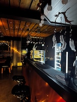 Uma área de bar com balcão, banquetas, luminárias pendentes e uma vitrine refrigerada.
