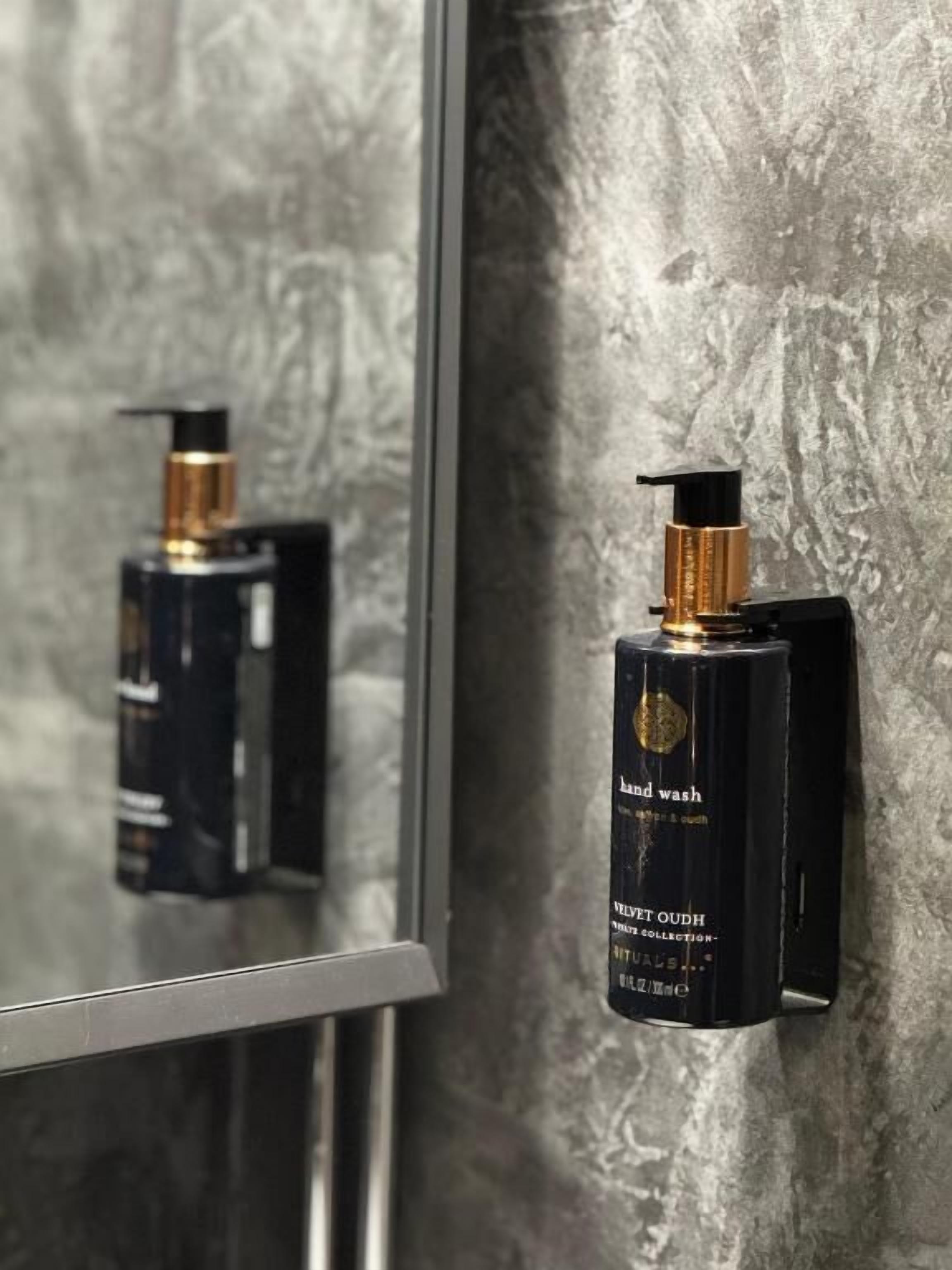A hand wash dispenser mounted on a textured wall, with a mirror reflecting its image.