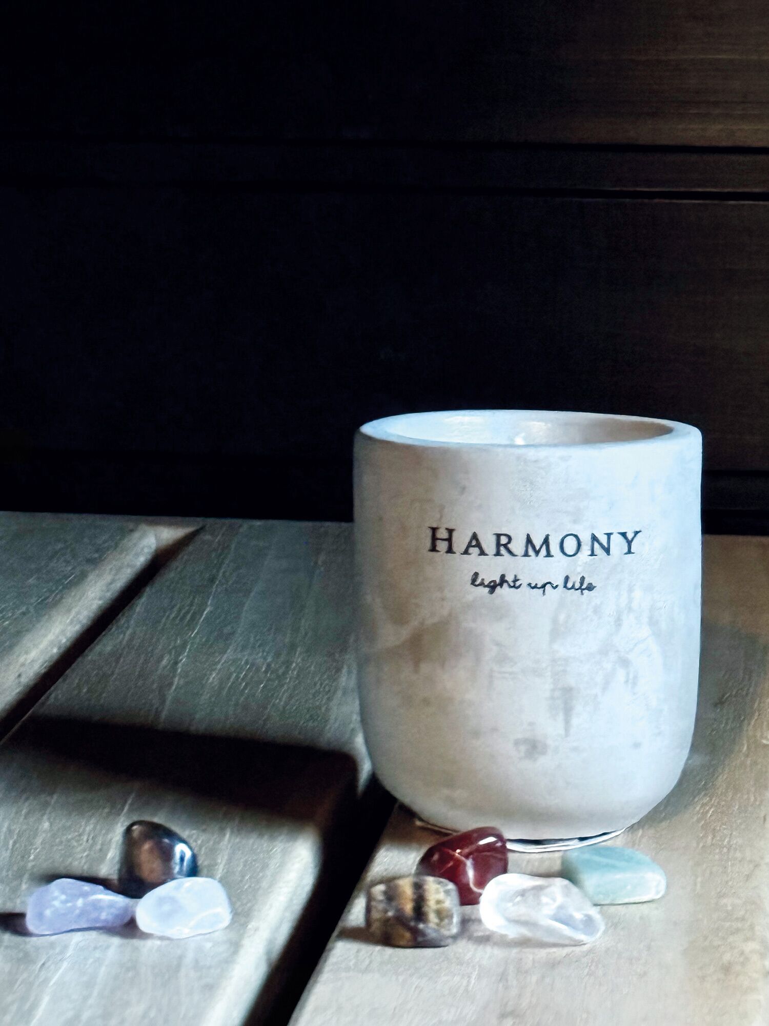 Ett ljus med texten ”HARMONI” och ”sprid ljus i livet”, placerat på en träyta med utspridda stenar.