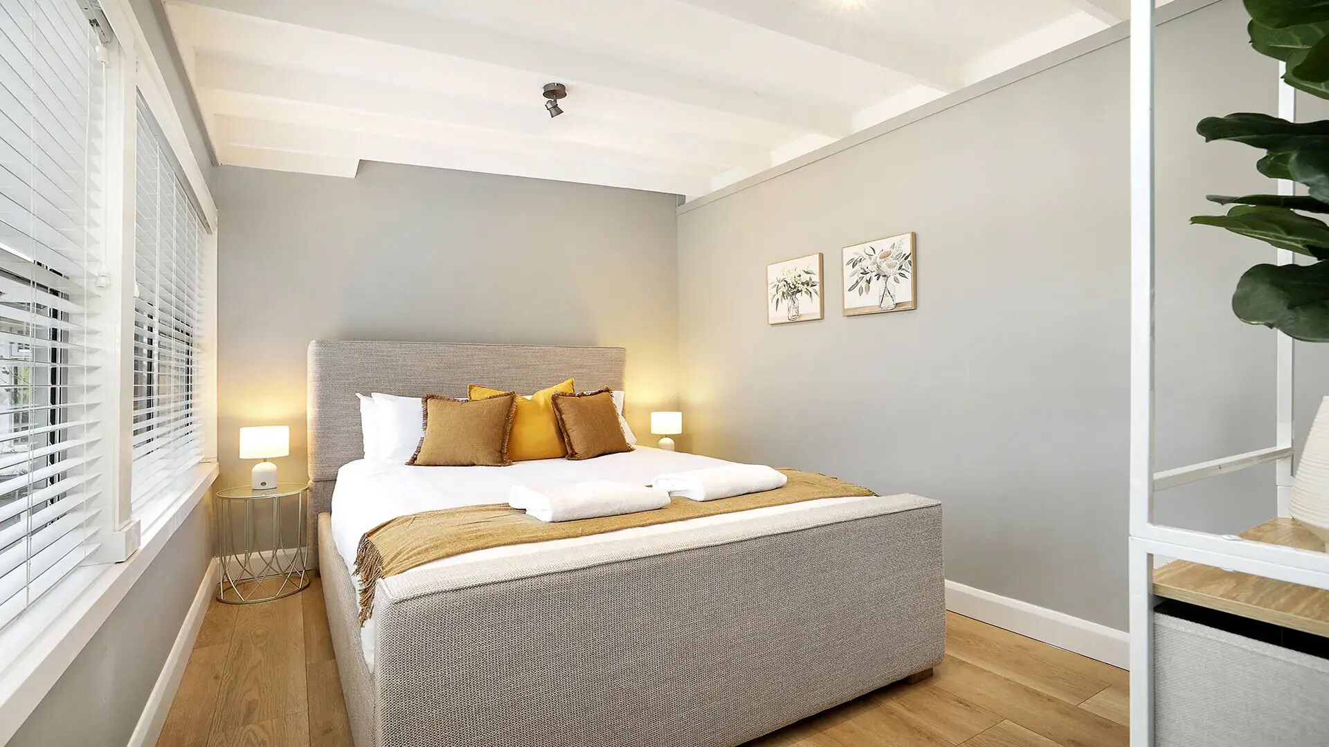 A bedroom with a bed, bedside lamps, a window with blinds, and a plant.