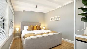 A bedroom with a bed, bedside lamps, a window with blinds, and a plant.