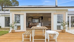 Een modern huis met een houten terras, een eethoek en een keuken die door de open deur zichtbaar is.
