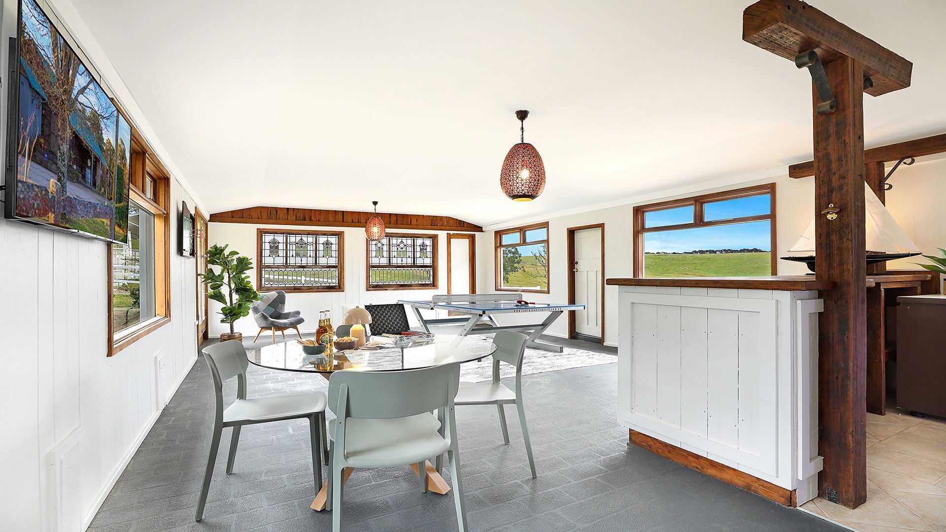A spacious room with a round table, chairs, a ping pong table, and large windows offering views of the outdoors.