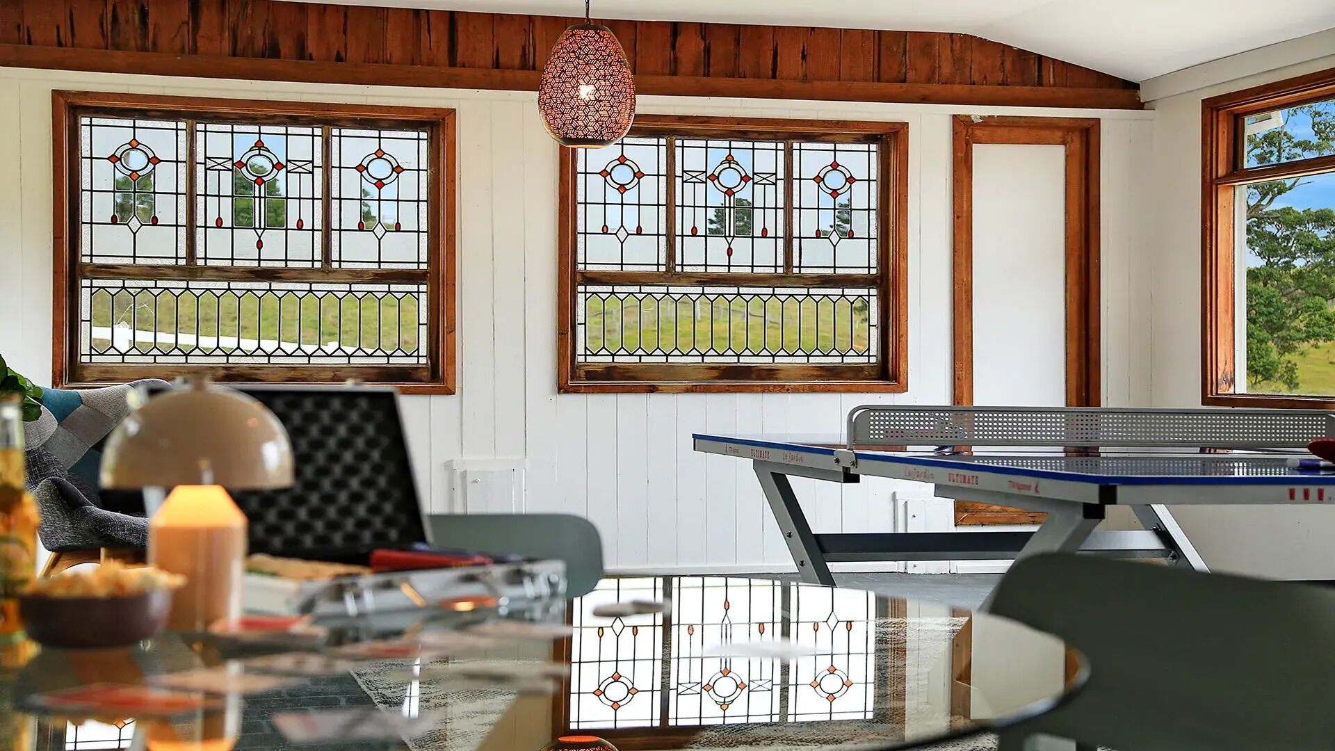 Una stanza con tavolo da ping pong, tavolo in vetro e una sedia. Sono presenti due finestre con vetrate decorate e un lampadario.